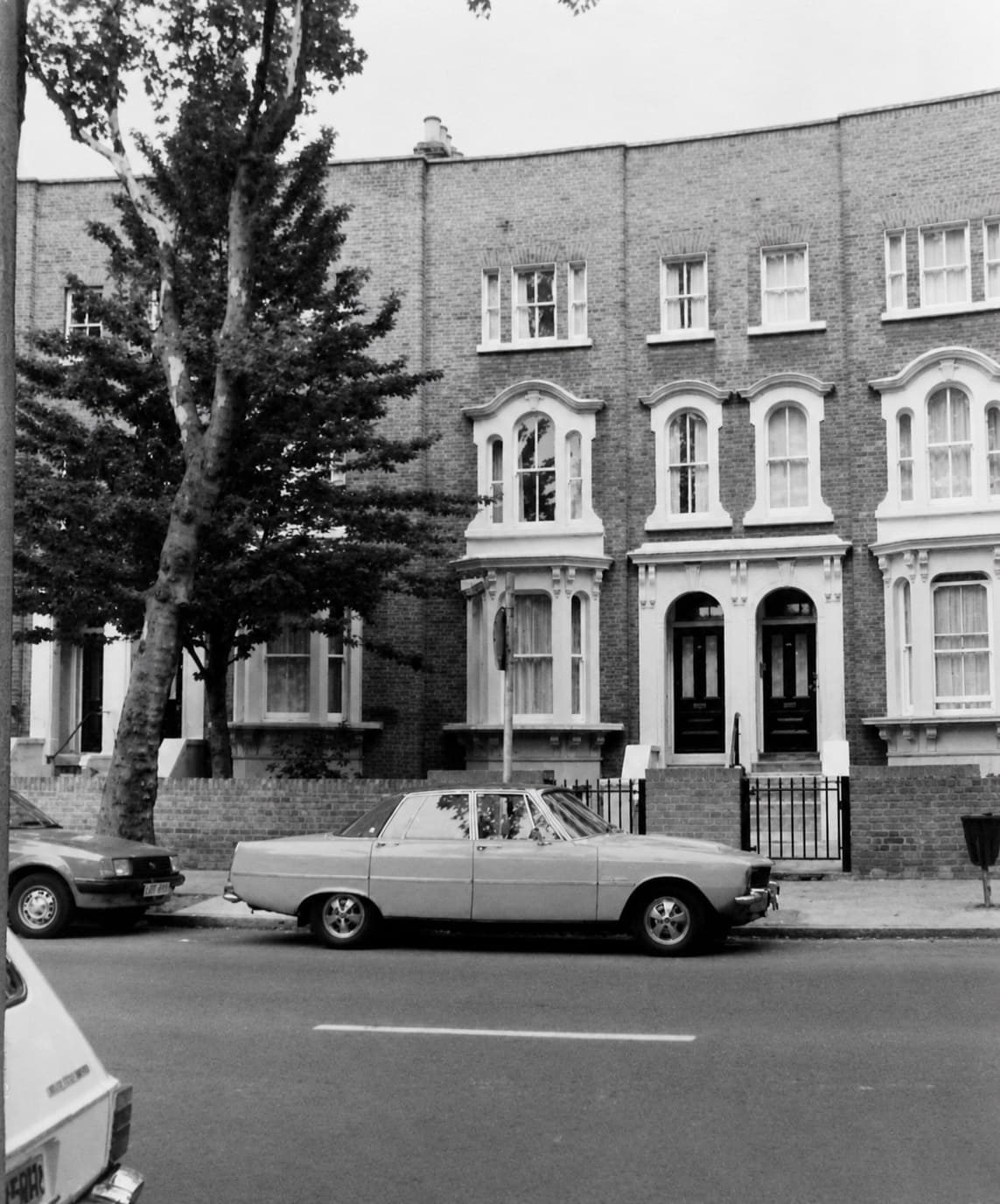 Black-and-white photograph of a Victorian terrace in Hackney with bay windows and arched entrances, a Rover saloon parked at the kerbside, photographed in the early 1980s.